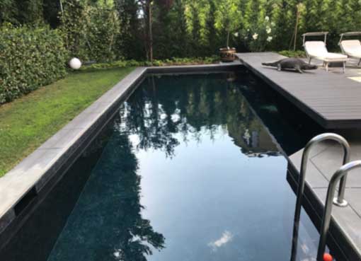Piscina interrata nera con bordo in pietra e area relax con lettini in giardino.
