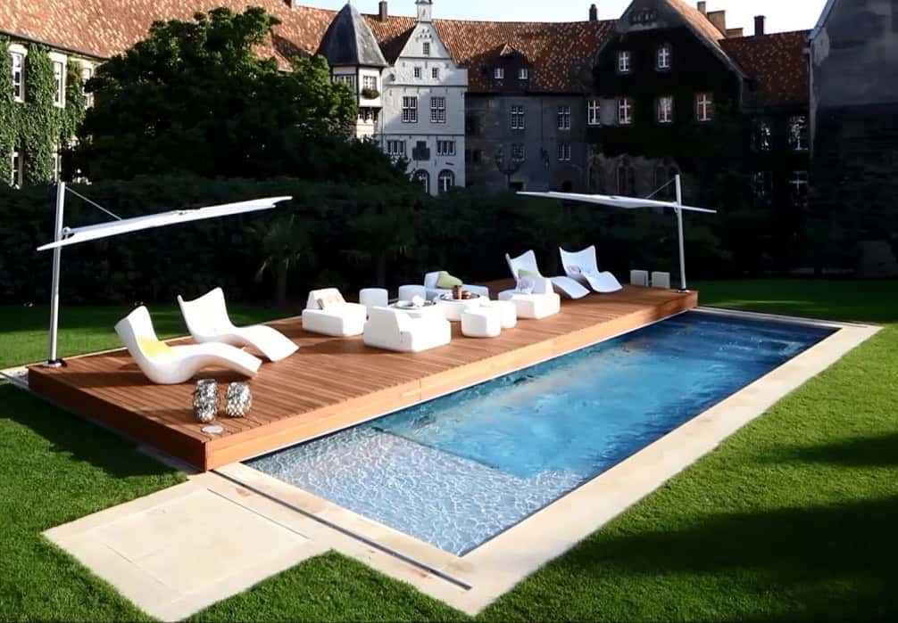 Piscina privata con zona relax in legno, chaise longue moderne e ombrelloni, ambiente outdoor, giardino curato, case storiche sullo sfondo, materiali di alta qualità, colori naturali e arredi eleganti.