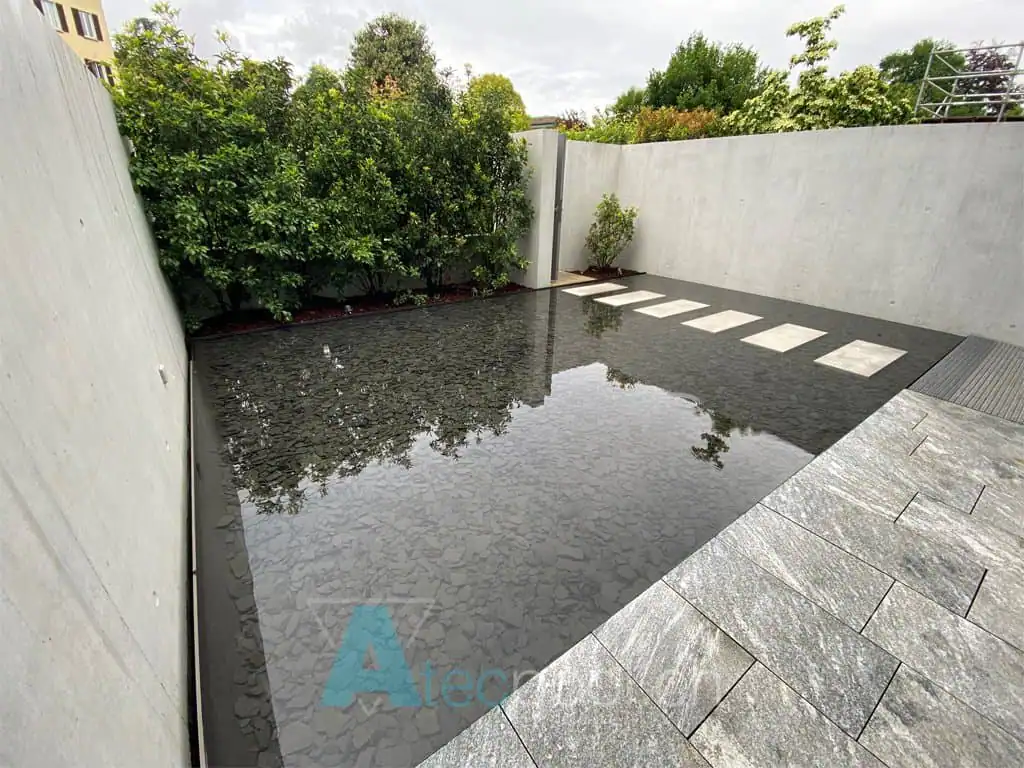 Piscina quadrata con finitura scura riflettente, design minimale, lastre sospese e cornice in cemento — realizzata da Atec Pool in un giardino moderno
